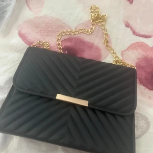 Svart quiltad axelväska med guldkedja - Stilren svart axelväska med quiltat mönster och snygg guldfärgad kedja som axelrem. Väskan har ett elegant guldfärgat spänne framtill och magnetlås. Perfekt storlek för det viktigaste och ger en lyxig känsla till din outfit.