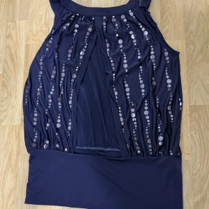 Lila  linne med paljetter - Snygg lila linne med glittriga paljettdetaljer i vågiga rader framtill. Linet  är ärmlös och har en bred kant nertill som ger en cool siluett. Perfekt för dig som vill sticka ut med lite extra bling.