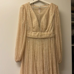 Champagnefärgad glittrig långklänning - Magisk långklänning i champagnefärg med glittriga paljetter över hela tyget. Klänningen har v-ringning, markerad midja och långa, luftiga ärmar. Materialet är lätt och skimrande, perfekt för att sticka ut. Snyggt fall och romantisk känsla.och klänningen har även ett lite längre släp där bak vilket ger en lyxig känsla på klänningen. 