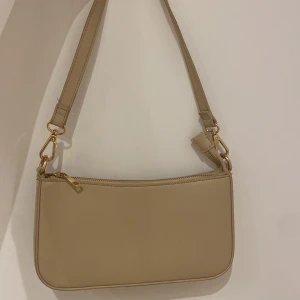 Beige axelväska i skinn med gulddetaljer - Stilren beige axelväska i skinn med guldfärgade metalldetaljer. Väskan har en enkel, rektangulär form, dragkedja upptill och en extra ficka med dragkedja på baksidan. Perfekt för dig som gillar minimalistisk och clean stil.