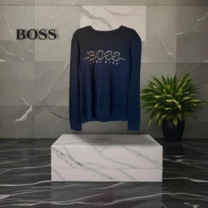 Svart slim fit tröja från Hugo Boss - Svart långärmad tröja från Hugo Boss med broderad logga i guld på bröstet. Slim fit-modell med rund halsringning och ribbade muddar. Perfekt för dig som gillar stilrena och klassiska plagg med en exklusiv touch.