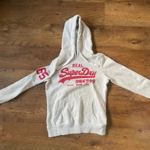 Grå hoodie från Superdry - Snygg ljusgrå hoodie från Superdry med rött tryck på bröstet och siffran 65 på ärmen. Tröjan har huva med snörning och en stor magficka. Perfekt för chill dagar eller när du vill ha en sportig look.