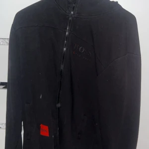 Svart hoodie från Hugo Boss - Svart hoodie från Hugo Boss med dragkedja framtill och diskret logga på bröstet. Tröjan har huva och fickor på sidorna samt en röd detalj med Hugo Boss-märke nedtill. Perfekt för en chill och stilren look.