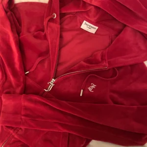 Röd velour hoodie från Juicy Couture - Juicy Couture hoodie i röd velour med glittrande strasstenar på ryggen och JC-detalj framtill. Tröjan har dragkedja, huva med snörning och långa ärmar. Bra skick . Storlek S