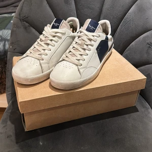Pepe Jeans vita sneakers med blå detalj - Snygga vita sneakers från Pepe Jeans med blå detalj på sidan och svart mocka vid hälen. Skorna har snörning, platt sula och cool 1973-detalj bak. Tillverkade i skinn och mocka för en stilren look. Perfekta för dig som gillar klassisk streetstyle.