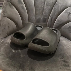 Svarta Adidas Yeezy Slides - Svarta Adidas Yeezy Slides med chunky design och öppen tå. Tillverkade i syntetmaterial med räfflad sula för extra grepp. Perfekta för chill dagar och enkel att slänga på sig. Diskret Adidas-logga på innersulan.