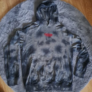 Stranger Things hoodie från H&M, M - Säljer en grå och svart tie-dye hoodie från H&M x Stranger Things. Framsidan har röd Stranger Things-logga och baksidan har ett stort tryck med karaktärer och röd effekt. Hoodien har känguruficka och huva, relaxed fit. Perfekt för dig som älskar serien!