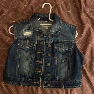 Mörkblå jeansväst från Springfield - Snygg mörkblå jeansväst från Springfield med klassisk krage, knappar framtill och två bröstfickor med lock. Västen har slitna detaljer och kontrastsömmar som ger en cool look. Perfekt att slänga över en t-shirt eller hoodie för extra edge.