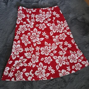 Röd blommig kjol från Clockhouse - Röd A-linjeformad kjol från Clockhouse med stora vita blommor. Kjolen är i 100% bomull och har en diskret dragkedja i sidan. Perfekt för dig som vill sticka ut med ett färgstarkt och somrigt plagg. Den saknar en knapp vid toppen av dragkedjan, men det är inget som påverkar hur kjolen sitter och syns inte när den är på.