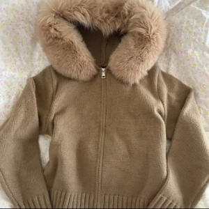 Beige stickad hoodie med fuskpäls - Supermysig beige stickad hoodie med dragkedja och stor huva kantad av fluffig fuskpäls. Tröjan har ribbade muddar och midja, perfekt för kalla dagar. Snygg och chill passform som funkar till jeans eller leggings.