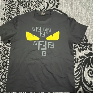 Svart t-shirt med gul ögonprint - Svart t-shirt i bomull med coolt tryck framtill: två gula ögon och flera grå F-mönster. Klassisk rund hals och korta ärmar. Perfekt för dig som vill sticka ut med en edgy vibe.