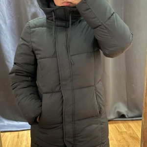 Svart lång pufferjacka från H&M XS - Svart lång pufferjacka från H&M i storlek XS. Jackan har hög krage med snörning, dragkedja och tryckknappar framtill samt två sidofickor. Perfekt för kalla dagar med sin quiltade design och rejäla vaddering.