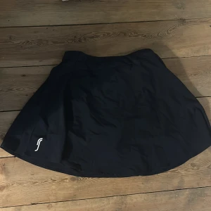 Svart sportig kortkjol från Swedemount - Svart sportig kortkjol från Swedemount med enkel design och liten vit logga framtill. Kjolen har en lätt utsvängd form och är tillverkad i ett syntetmaterial som passar perfekt för aktiva dagar.