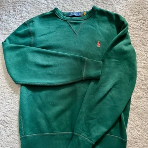 Mörkgrön sweatshirt från Polo Ralph Lauren - Mörkgrön sweatshirt från Polo Ralph Lauren med klassisk rund hals och broderad logga på bröstet. Tröjan har långa ärmar och är tillverkad i mjuk bomull, perfekt för en avslappnad och stilren look.