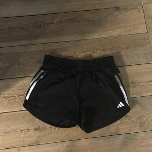 Svarta Adidas träningsshorts - Snygga svarta träningsshorts från Adidas med vita ränder på sidorna och elastisk midja. Perfekta för gymmet eller löprundan, med klassisk sportig look och lätt material som andas. Adidas-logga på ena benet för extra stilpoäng.