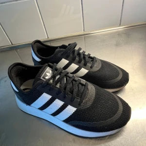 Svarta Adidas sneakers med vita ränder - Snygga svarta Adidas sneakers med klassiska vita ränder på sidorna och svart/vit sula. Ovandelen är i stickad syntet med snörning och rund tå. Perfekta för dig som gillar sportig och stilren look. ( lite skav vid ena tån ) 