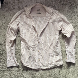 Randig skjorta från Jack & Jones XS - Snygg ljusbeige skjorta från Jack & Jones Essentials med vita vertikala ränder. Skjortan har klassisk krage, knäppning framtill och långa ärmar. Tillverkad i en skön mix av bomull och linne, perfekt för varma dagar.