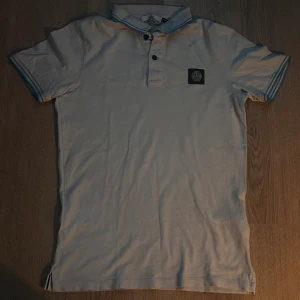 Vit Stone Island piké med blå detaljer - Vit pikétröja från Stone Island med blå ränder på krage och ärmslut. Klassisk passform, kortärmad och med svart logotyp-patch på bröstet. Tillverkad i bomull och har knappslå framtill. Perfekt för en clean och stilren look. Det finns små hål på den men man kan knappast se dom.