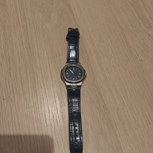 Blå klocka från Regal med silverboett - Stilren klocka från Regal med blått läderarmband och silverfärgad boett. Urtavlan är blå med diskreta ränder och vita markeringar. Klassisk design som passar till många stilar. Armbandet har krokodilpräglad yta för extra detalj.