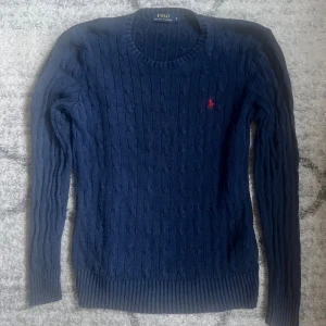 Marinblå stickad tröja Polo Ralph Lauren - Kabelstickad marinblå tröja från Polo Ralph Lauren med klassisk rund halsringning och röd broderad logga på bröstet. Långärmad modell i mjuk bomull som passar perfekt till jeans eller kjol.