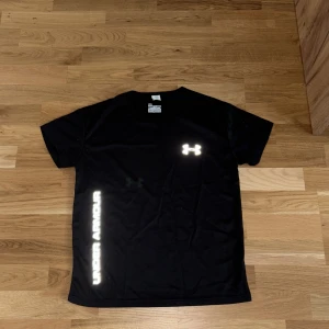 Svart Under Armour t-shirt  - Svart t-shirt från Under Armour med reflekterande logga på bröstet och text längs sidan. Den är tillverkadei lätt, ventilerande polyester som passar perfekt för träning. Klassisk rund hals och korta ärmar för en sportig look. Storleken är M men passar XS