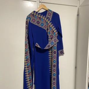 Blå palestinsk klänning  - En lång, kornblå palestinsk klänning med långa ärmar. Klänningen har färgstarka broderade detaljer i gult, lila och blått längs ena sidan, ärmslut och medföljande sjal. Perfekt för dig som vill sticka ut med unika mönster och färger. Det finns även en blå underklänning i samma färg. 