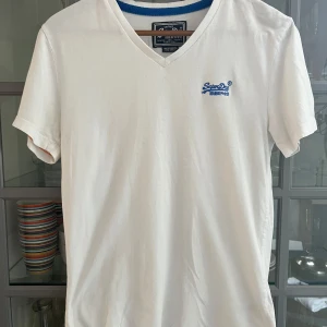 Vit v-ringad t-shirt från Superdry - Vit t-shirt från Superdry med v-ringning och blå broderad logga på bröstet. Klassisk passform och tillverkad i mjuk bomull. Perfekt basic-plagg med snygga detaljer och diskret branding.