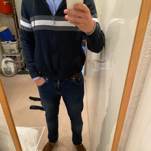 Marinblå hoodie från Massimo Dutti stickad - Snygg marinblå hoodie från Massimo Dutti med dragkedja och huva. Tröjan har två horisontella ränder i vitt och grått över bröstet och är tillverkad i mjuk bomull. Perfekt för en chill och stilren look.