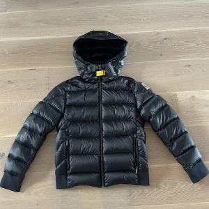 Svart dunjacka med huva från Parajumpers - Snygg svart dunjacka från Parajumpers med glansig finish, huva och ikonisk gul detalj vid halsen. Jackan har dragkedja framtill, två sidofickor med dragkedja och ribbade muddar. Perfekt för kalla dagar och riktigt stilren look.