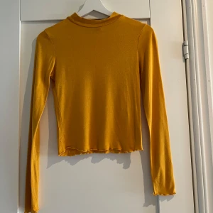 Gul långärmad polotopp från Gina Tricot - Säljer en snygg gul långärmad polotopp från Gina Tricot i storlek XS. Toppen har en tight passform, liten polokrage och vågiga kanter vid ärmslut och nederkant. Perfekt att styla med jeans eller kjol för en cool look.