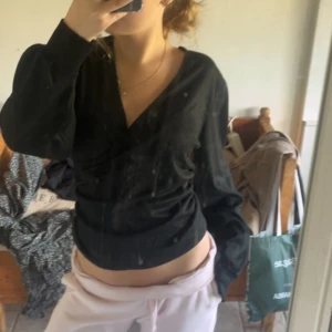 Svart omlottopp med lång ärm - Säljer en svart omlottopp med lång ärm och v-ringning. Snygg och figurnära passform som framhäver midjan. Perfekt att styla med jeans eller kjol för en clean look.