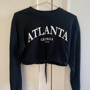 Svart croppad tröja från Bershka - Svart croppad sweatshirt från Bershka med vit text 'ATLANTA GEORGIA U.S.A.' framtill. Tröjan har långa ärmar, rund halsringning och snörning i nederkanten för en snygg passform. Perfekt för en chill och trendig look.