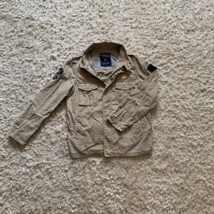 Beige overshirt från Massimo Dutti - Snygg beige overshirt från Massimo Dutti med coola patches på ärmarna och tryck på ryggen. Jackan har fyra fickor framtill, klassisk krage och stängs med knappar. Tillverkad i 100% bomull, perfekt för hösten. Hittar ingen storlek men skulle säga att den sitter som en S, tveka inte o skriva vid frågor!