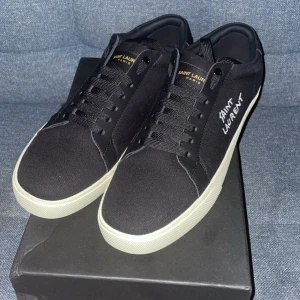 Svarta Saint Laurent sneakers - Snygga svarta sneakers från Saint Laurent med vit sula och broderad logga på sidan. Skorna har klassisk snörning och är tillverkade i textil med detaljer i skinn. Perfekta för dig som gillar stilrena och exklusiva sneakers med en modern vibe. Kvitto finns
