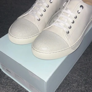 Vita sneakers från Lanvin - Snygga vita sneakers från Lanvin i lyxigt skinn med texturerad tå och klassisk snörning. Skorna har rund tå, platt sula och diskreta silverfärgade metalldetaljer. Perfekta för dig som gillar stilrena och exklusiva sneakers med en clean look. 100% äkta VERIFIERADE MED VINTED bevis medföljer. Om du har några fråga bara skriv!