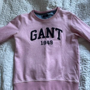 Rosa sweatshirt från GANT med tryck - Rosa sweatshirt från GANT med svart broderat logotyp-tryck och årtalet 1949 på bröstet. Tröjan har rund halsringning, ribbade muddar och är tillverkad i mjuk bomullsmix. Perfekt för en chill och sportig look.