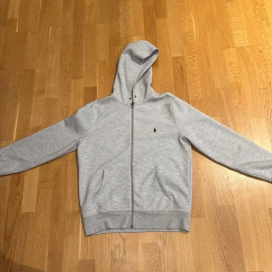 Grå hoodie från Polo Ralph Lauren - Snygg ljusgrå hoodie från Polo Ralph Lauren med dragkedja framtill och klassisk broderad logga på bröstet. Tröjan har huva, två fickor och är tillverkad i mjuk bomullsblandning. Perfekt för chill dagar eller när du vill ha en clean look. Snöre saknas annars bra skick
