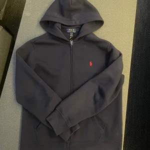 Mörkblå zip up hoodie från Polo Ralph Lauren - Mörkblå zip up hoodie från Polo Ralph Lauren med dragkedja och huva. Klassisk röd broderad logga på bröstet och känguruficka framtill. Tillverkad i mjuk bomullsmix, perfekt för chill dagar. Snygg och enkel att matcha med allt. Aldrig använt innan och är i mycket bra skick! 