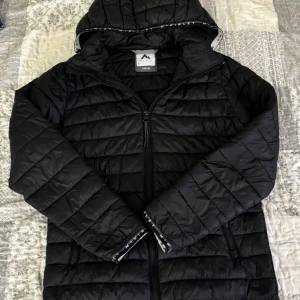 Svart pufferjacka från McKinley - Svart pufferjacka från McKinley med huva och dragkedja. Jackan är quiltad och har två sidofickor med dragkedja. Yttertyg och foder i nylon, fyllning i polyester. Perfekt för kyliga dagar och har en clean, sportig look.