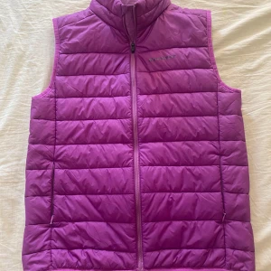 Lila dunväst från Everest - Lila dunväst från Everest med quiltad design och hög krage. Västen har två fickor med dragkedja och hel dragkedja framtill. Perfekt för lager-på-lager och ger en sportig vibe. Snygg och funktionell för kyligare dagar.