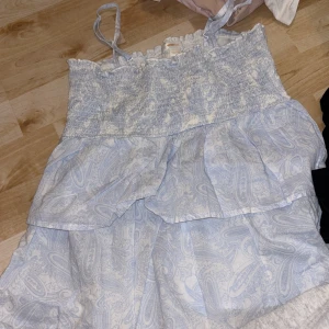 Ljusblått linne från Gina Tricot - Supersöt ljusblå linne från Gina Tricot med paisleymönster och smock upptill. Linnet har tunna axelband och volangdetaljer som ger en luftig och somrig känsla. Perfekt för varma dagar och enkel att styla med sneakers eller sandaler.