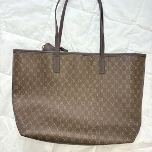 Brun shoppingväska från Don Donna - Stilren brun shoppingväska/Toteväska från Don Donna med klassiskt monogrammönster över hela väskan. Väskan har långa handtag, guldfärgade metalldetaljer och är tillverkad i syntetmaterial med skinnliknande känsla. Perfekt storlek för att få plats med allt du behöver till dagen. 