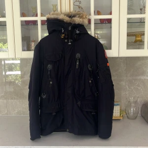 Svart vinterjacka Parajumpers Anchorage - Snygg svart Parajumpers Anchorage vinterjacka med päls på huvan, flera dragkedjor och fickor samt patch på ärmen. Jackan har hög krage, justerbar huva och coola detaljer som orange flärp och robusta knappar. Perfekt för kalla dagar.