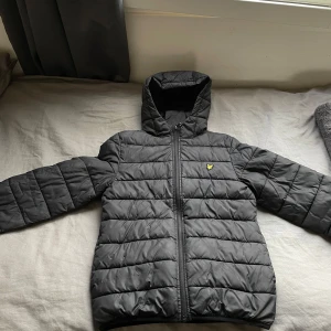 Svart pufferjacka Lyle & Scott Junior - Svart pufferjacka från Lyle & Scott Junior med huva och gul broderad logga på bröstet. Jackan har quiltad design, dragkedja framtill och är långärmad. Perfekt för kalla dagar och har en clean, modern look. Den är lite smutsig men annars i bra skick
