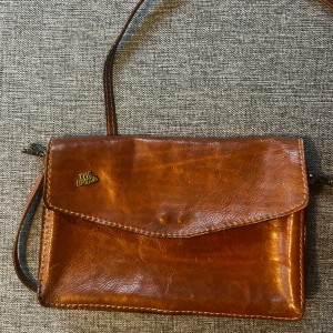 Brun axelväska i skinn från The Bridge - Snygg brun axelväska från The Bridge i äkta skinn med klassisk design. Väskan har lock med magnetknäppning, flera fack inuti och en smidig axelrem. Perfekt för dig som gillar stilrena och tidlösa accessoarer med italiensk känsla.