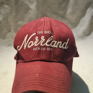 Vinröd keps The Great Norrland - Vinröd keps från Sqrtn Company med broderad text 'The Great Norrland' framtill och 'Sqrtn & Company' baktill. Kepsen har en broderad hjortron-detalj på sidan och justerbar snapback. Tillverkad i bomull och har klassisk böjd skärm.