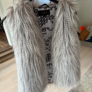 Ljusgrå pälsväst från Vila - Säljer en fluffig och trendig ljusgrå pälsväst från Vila. Västen har lång, mjuk fuskpäls och är öppen framtill utan knäppning. 💓