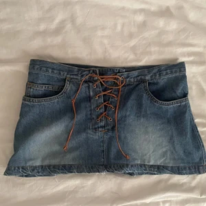 Mini jeans kjol med snörning - Säljer en blå jeanskjol med snygg snörning framtill och klassiska fickor både fram och bak. Kjolen är kort och har en cool, avslappnad vibe med låg midja. Perfekt för dig som gillar denim och vill ha något trendigt i garderoben. Skulle säga att den passar xxs-s då man kan justera den i midjan💕