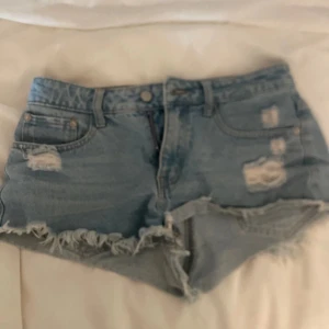 Blå slitna jeansshorts med fransar - Säljer ett par blå jeansshorts med slitna detaljer och fransig kant. Shortsen har klassisk femficksmodell och dragkedja framtill. Perfekta för en avslappnad och trendig look i sommar.