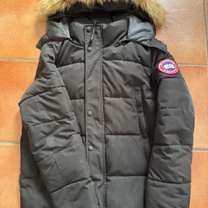 Svart dunjacka från Canada Goose S - Säljer en svart dunjacka från Canada Goose med klassisk patch på ärmen och avtagbar huva med fuskpäls. Jackan är quiltad, har dragkedja och knappar framtill samt stora fickor. Perfekt för kalla vinterdagar och riktigt snygg streetstil.